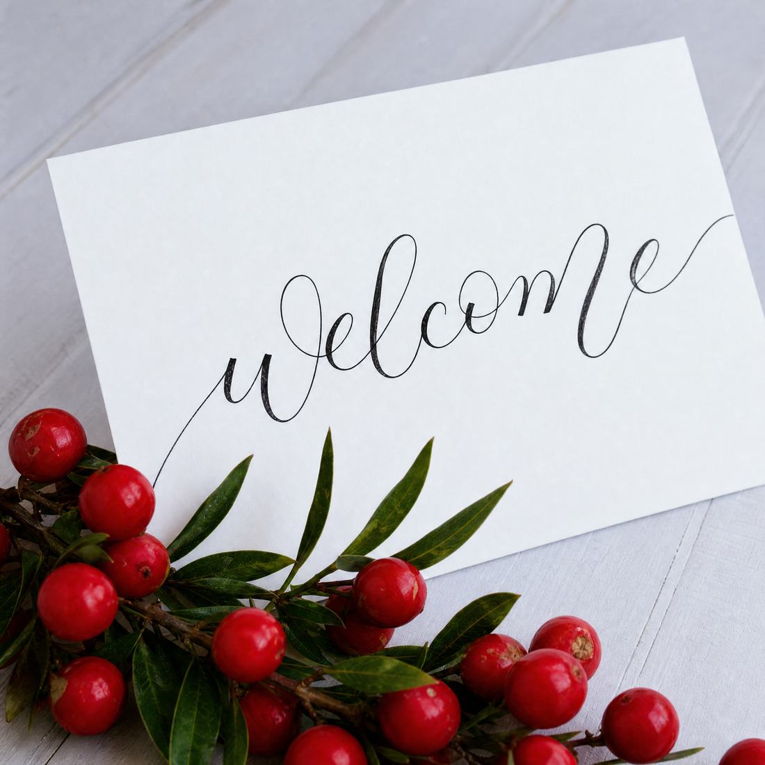 The word welcome in flourished copperplate calligraphy on a white card beside red berries