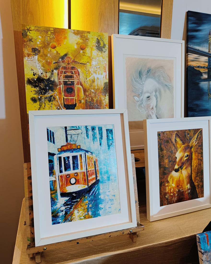 Framed paintings by Vinda Shinde on exhibition easels, including a vintage tram and a deer in amber tones