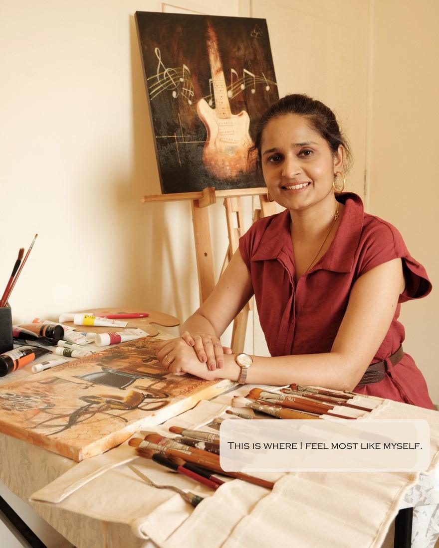 Vinda Shinde at her easel with brushes and paints, a guitar painting behind her